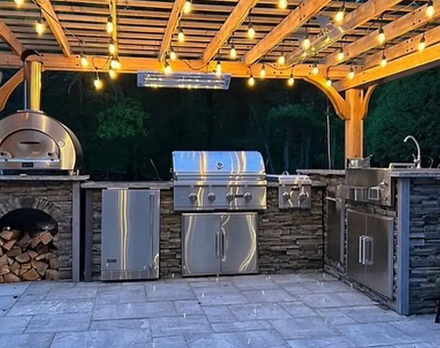 Outdoor kitchen with grill and pizza oven under a lit cedar pergola